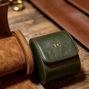 May include: A dark green leather watch roll with a gold embossed monogram TC on the front flap. The interior features a soft brown suede lining with a cylindrical cushion for watch storage, showcasing a smooth texture and stitched edges.