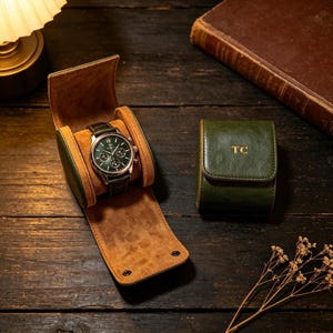 Könnte beinhalten: Ein dunkelgrünes Uhrenetui mit beigefarbenem Innenfutter, das eine silberne Chronographenuhr mit grünem Zifferblatt enthält. Daneben steht ein geschlossenes, passendes grünes Uhrenetui mit den goldgeprägten Initialen TC auf der Vorderklappe.