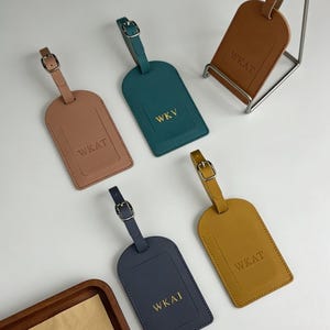 Peut inclure: Plusieurs étiquettes de bagages en cuir de différentes couleurs, dont beige, sarcelle, marine et jaune moutarde. Chaque étiquette comporte une sangle à boucle et du texte en relief, avec les lettres "WKV" ou "WKAT" visibles.