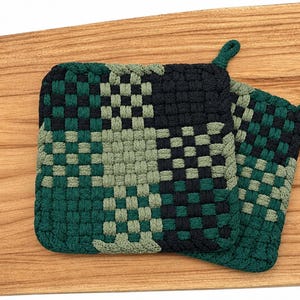 May include: Two square potholders featuring a woven checkerboard pattern in shades of dark green, light green, and black. Each potholder is constructed from thick, interlocking fabric loops and includes a small fabric loop at the corner for hanging.