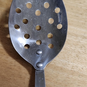 Op de afbeelding: Een roestvrijstalen sleef met een lange steel en een ovale kop. De kop heeft meerdere ronde gaten om vloeistoffen af te voeren. De lepel is ontworpen voor gebruik bij het koken en bereiden van voedsel.