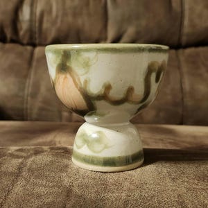Könnte beinhalten: Ein weißer Eierbecher aus Keramik mit einem grünen und braunen Blumendesign. Der Becher hat einen Sockel.