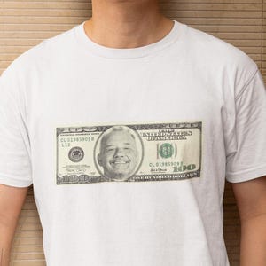 May include: White t-shirt featuring a graphic of a $100 bill. The shirt is worn by a person in light-colored pants. The background is a textured, light brown wall.