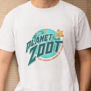 May include: A white t-shirt featuring the text "PLANET ZOOT" in teal and green, with a star and planet graphic. The phrase "THE SECOND COMING OF ZOOT" is below the main text. The shirt is worn by a person.