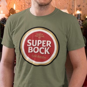 May include: Olive green t-shirt featuring a circular logo with the words "SUPER BOCK" in bold white letters on a red background. The logo also includes text in Portuguese. The shirt is short-sleeved.