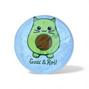 Peut inclure: Un bouton rond bleu clair avec un motif de chat avocat de dessin animé. L'avocat chat est vert avec un noyau marron et des moustaches noires. Le texte "Guac & Roll" est imprimé en noir.