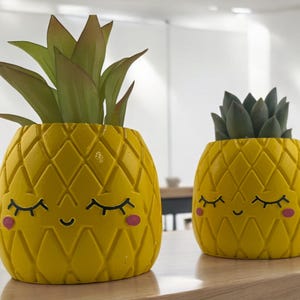 May include: Two yellow pineapple-shaped planters with smiling faces and closed eyes. The planters are made of ceramic and have a textured surface. The larger planter has a green leafy plant inside, while the smaller planter has a green succulent inside.