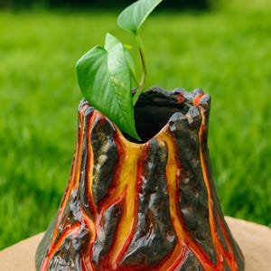 Peut inclure: Un pot de fleurs en céramique en forme de volcan avec une plante. Le pot est peint avec un motif texturé en noir, rouge et jaune, ressemblant à de la lave. La plante a de grandes feuilles vertes.