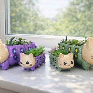 May include: Two sets of ceramic cat-shaped planters, each featuring a large and small version. One set is purple with blue and dark purple circular patterns, and the other is sage green with yellow and dark green circular patterns on the body.