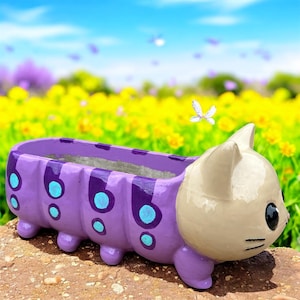 May include: A whimsical, purple planter shaped like a caterpillar with a cat's head. The planter has a light beige cat face, turquoise spots, and dark purple stripes. The planter is sitting on a stone surface with a blurred background of yellow flowers and a blue sky.