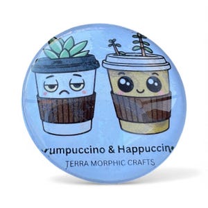 May include: A light blue, round button featuring two cartoon coffee cups. One cup is labeled "Yumpuccino" and has a sad face and a succulent on top. The other cup is labeled "Happuccino" with a happy face and a plant. The text "Terra Morphic Crafts" is also present.