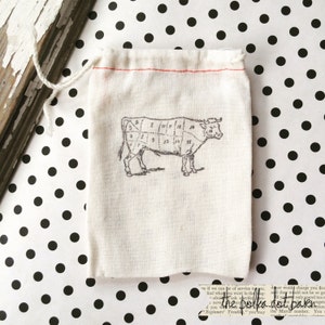 May include: A small white cotton drawstring bag with a red drawstring. The bag has a black and white illustration of a cow with numbered cuts of meat printed on it. The bag is sitting on a white background with black polka dots.