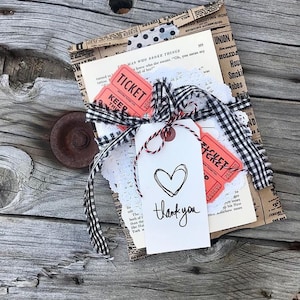 May include: A gift wrapped in brown paper with black and white checkered ribbon, white lace, and two red tickets. A white tag with a hand-drawn heart and the text "thank you" is attached to the package.