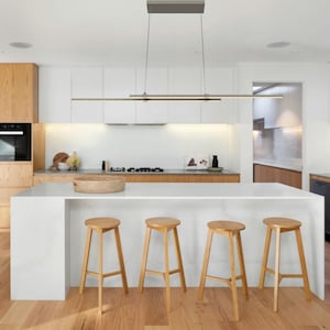 May include: Modern kitchen with light wood cabinetry, white countertops, and a large island with four wooden stools. A linear pendant light hangs above the island. The flooring is light-colored wood.