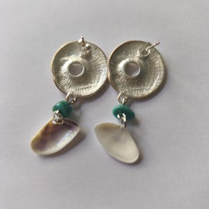 Peut inclure: Une paire de boucles d'oreilles &agrave; clous argent&eacute;es compos&eacute;es d'un disque m&eacute;tallique circulaire avec un motif radial textur&eacute;. Chaque boucle est orn&eacute;e d'une petite perle lisse couleur turquoise et d'un pendentif en coquillage naturel blanc cass&eacute;.