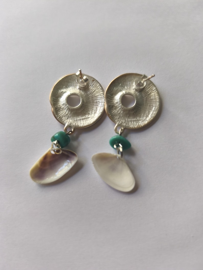 Peut inclure: Une paire de boucles d'oreilles &agrave; clous argent&eacute;es compos&eacute;es d'un disque m&eacute;tallique circulaire avec un motif radial textur&eacute;. Chaque boucle est orn&eacute;e d'une petite perle lisse couleur turquoise et d'un pendentif en coquillage naturel blanc cass&eacute;.