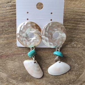 Puede incluir: Un par de pendientes colgantes con un disco de metal plateado martillado con un recorte central. Debajo de cada disco cuelga una pequeña cuenta de color turquesa brillante seguida de un dije de concha marina de color blanco roto en forma de abanico.