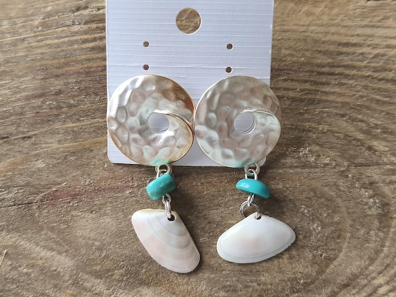 Peut inclure: Une paire de boucles d'oreilles pendantes compos&eacute;e d'un disque en m&eacute;tal argent&eacute; martel&eacute; avec une d&eacute;coupe centrale. Sous chaque disque pend une petite perle turquoise vive suivie d'une breloque en coquillage blanc cass&eacute; en forme d'&eacute;ventail.