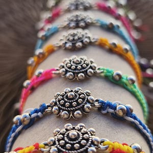May include: A collection of bracelets featuring silver-toned metal flower charms with intricate beaded details. Each bracelet is crafted from colorful braided cord in shades of yellow, blue, pink, green, and orange, accented with small silver beads.