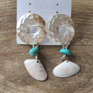 Peut inclure: Une paire de boucles d'oreilles pendantes compos&eacute;es d'un disque en m&eacute;tal argent&eacute; martel&eacute; avec un trou central, d'une petite perle turquoise et d'un coquillage blanc suspendu &agrave; un anneau en m&eacute;tal argent&eacute;.