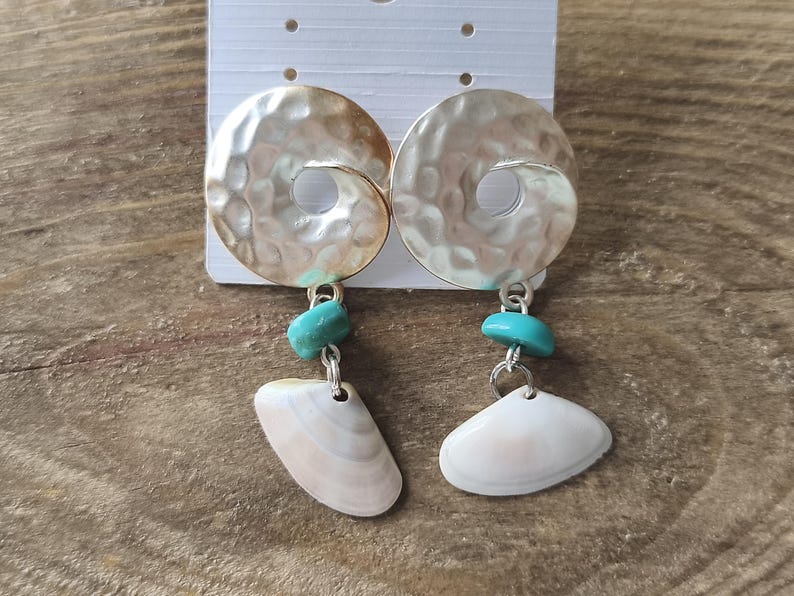 Peut inclure: Une paire de boucles d'oreilles pendantes compos&eacute;es d'un disque en m&eacute;tal argent&eacute; martel&eacute; avec un trou central, d'une petite perle turquoise et d'un coquillage blanc suspendu &agrave; un anneau en m&eacute;tal argent&eacute;.