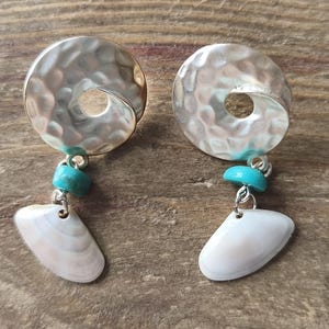 Peut inclure: Une paire de boucles d'oreilles pendantes compos&eacute;e d'un disque en m&eacute;tal argent&eacute; &agrave; la texture martel&eacute;e. Chaque boucle comprend une petite perle cylindrique de couleur turquoise et un coquillage blanc cass&eacute; lisse suspendu au disque m&eacute;tallique.