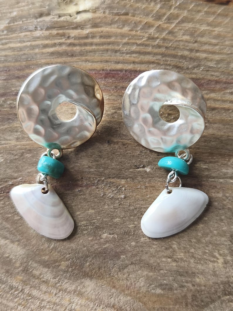 Peut inclure: Une paire de boucles d'oreilles pendantes compos&eacute;e d'un disque en m&eacute;tal argent&eacute; &agrave; la texture martel&eacute;e. Chaque boucle comprend une petite perle cylindrique de couleur turquoise et un coquillage blanc cass&eacute; lisse suspendu au disque m&eacute;tallique.