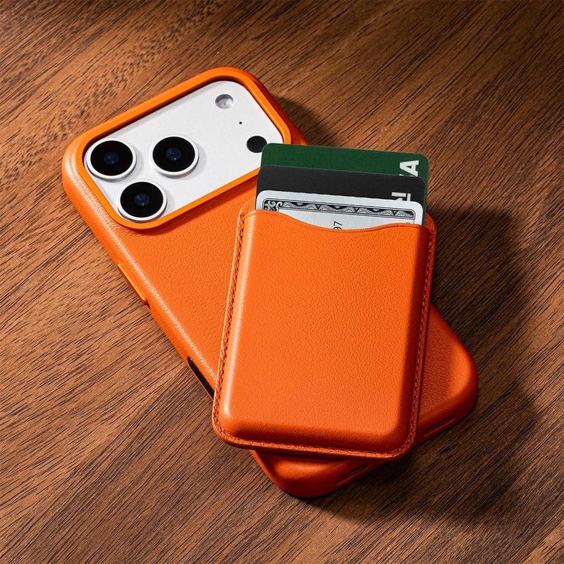 May include: Orange leather phone case with a card holder attached. The case is on a wooden surface. The card holder contains several cards. The phone case has a white camera section with three lenses.