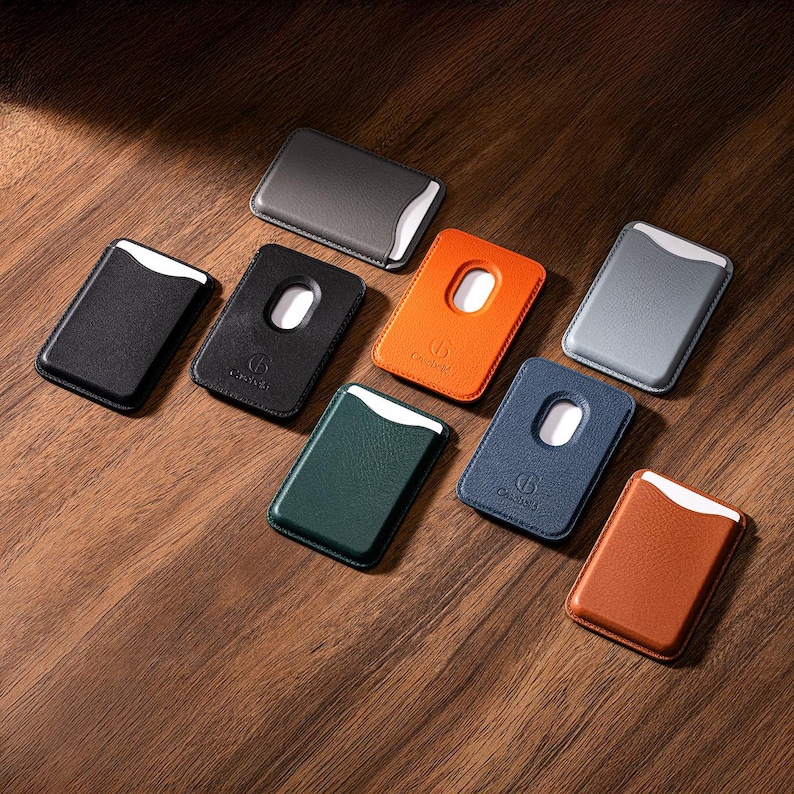 May include: A collection of leather cardholders in various colors, including black, gray, orange, green, and brown, arranged on a wooden surface. Each cardholder features a white accent and a unique design element, with some having a cutout.