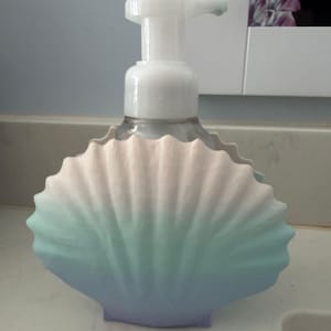 May include: A clear plastic soap dispenser with a white pump mechanism, encased in a decorative shell-shaped holder. The holder features a smooth gradient finish that transitions from a soft pale pink at the top to teal and light purple at the base.