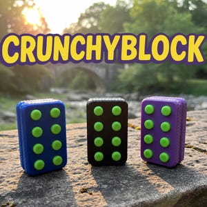 May include: Three rectangular fidget toys in blue, black, and purple, each with eight green circular buttons. The word "CRUNCHYBLOCK" is displayed in yellow and purple above the toys. The toys are on a stone surface with a blurred background of a river and trees.