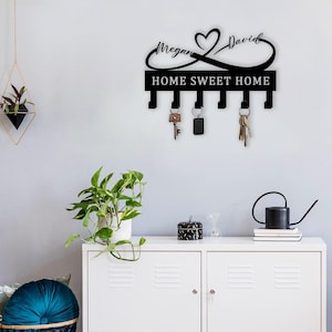 Pode incluir: Porta-chaves de metal preto com um símbolo de infinito e as palavras "Home Sweet Home". O símbolo de infinito tem um coração no meio e os nomes "Megan" e "David" escritos de cada lado. O porta-chaves tem cinco ganchos.
