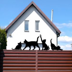 May include: Black metal cat silhouettes in different poses, mounted on a brown fence. One cat is seated, another is walking, a third is reaching for a butterfly, and the last is stretching. A white house with a brown roof is in the background.