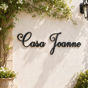 May include: A black metal wall sign featuring the cursive text "Casa Joanne" mounted on a light-colored stucco wall. The sign is positioned between green climbing vines and a black outdoor light fixture, creating a clear contrast in color.