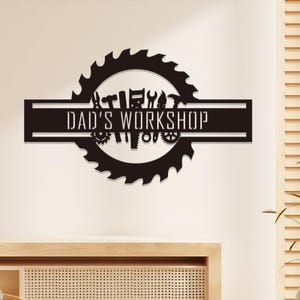 May include: A black metal sign with the text "DAD'S WORKSHOP" in white. The sign features a circular saw blade design with various tool silhouettes, including hammers, wrenches, and gears, creating a workshop theme.