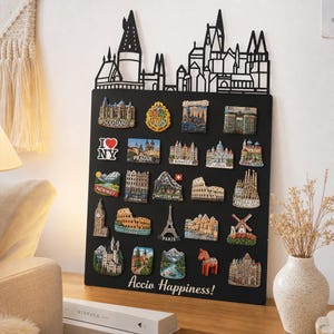 May include: A black rectangular magnetic display board featuring a laser-cut silhouette of a castle skyline at the top. The board is covered in various colorful travel souvenir magnets from locations like Scotland, Prague, and Paris, with the text Accio Happiness! printed in white at the bottom.