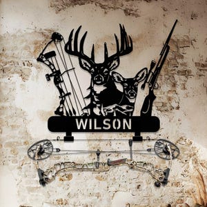 Peut inclure: Décoration murale en métal noir représentant une silhouette de cerf avec des bois, un fusil et un arc et des flèches. Le nom "WILSON" est affiché sur une bannière en dessous. Un deuxième arc et des flèches sont montés en dessous.