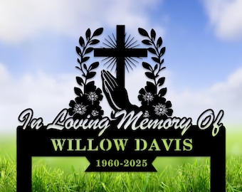 May include: Black metal memorial sign with a cross, praying hands, and floral accents. The sign reads "In Loving Memory Of" and "WILLOW DAVIS 1960-2025". The background shows a blue sky and green grass.