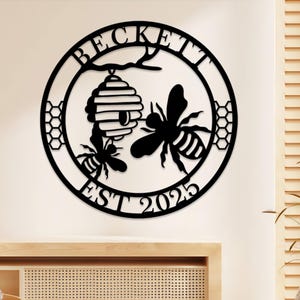 May include: Black metal wall art featuring a circular design with the name "BECKETT" at the top and "EST 2025" at the bottom. The design includes a beehive, two bees, and honeycomb patterns, perfect for home decor.