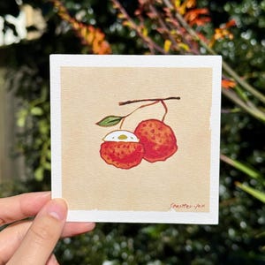 May include: A square watercolor painting of two red lychees with a brown stem and green leaf. One lychee is cut open, revealing white flesh and a brown seed. The painting is on a beige background and signed "Seastter-fox".