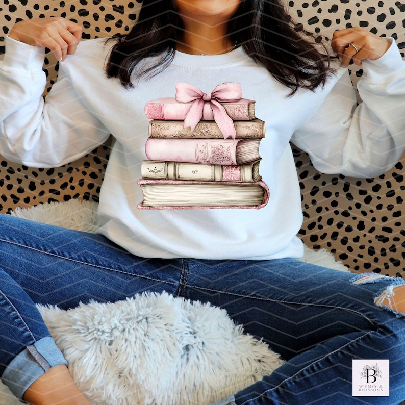 May include: A white sweatshirt with a stack of books tied with a pink ribbon. The books are illustrated with watercolor and have a vintage aesthetic.