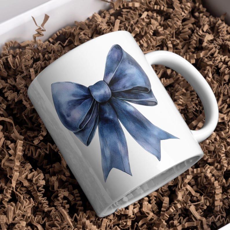 May include: A white ceramic mug with a blue watercolor bow design. The mug has a handle and is sitting on a brown paper surface.