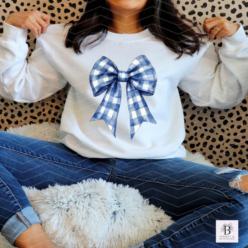 May include: A white sweatshirt featuring a blue and white gingham bow design. The bow is large and positioned centrally on the front of the sweatshirt.