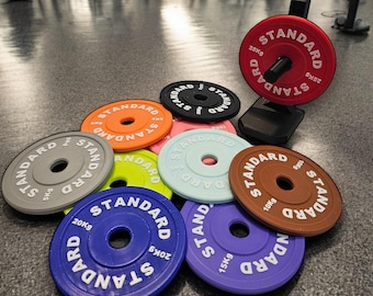 May include: A collection of ten weight plates in various colors, including gray, green, pink, blue, purple, and brown. Each plate has the word "STANDARD" printed on it, along with the weight in kilograms. The plates are arranged in a circle on a gray floor.