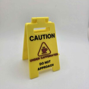 May include: Yellow plastic caution sign with black text that reads "CAUTION UNDER CAFFEINATED DO NOT APPROACH." The sign features a graphic of a coffee cup with steam. The sign is folded in a triangular shape.