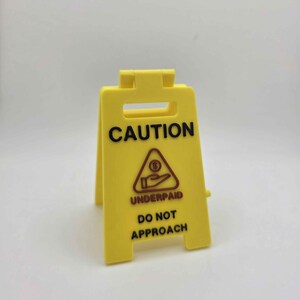 May include: A yellow plastic caution sign with the word "CAUTION" in black at the top. Below, a red triangle with a hand receiving money and the words "UNDERPAID DO NOT APPROACH" are printed in black.