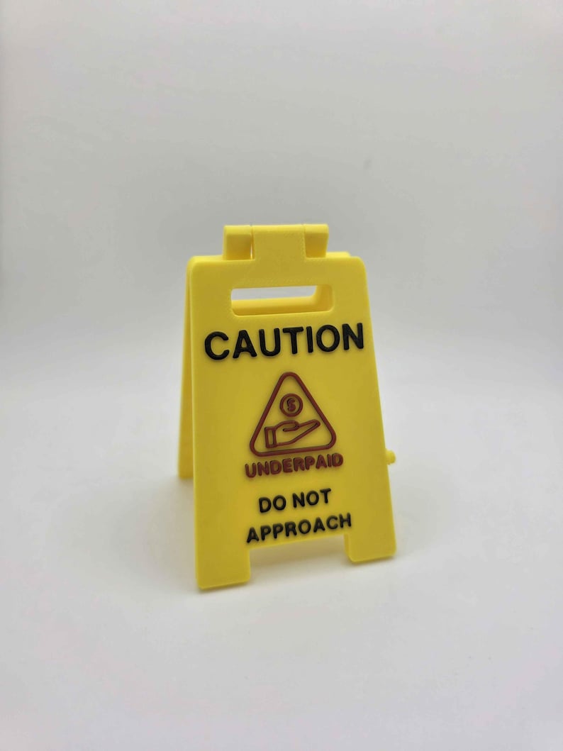 May include: A yellow plastic caution sign with the word "CAUTION" in black at the top. Below, a red triangle with a hand receiving money and the words "UNDERPAID DO NOT APPROACH" are printed in black.
