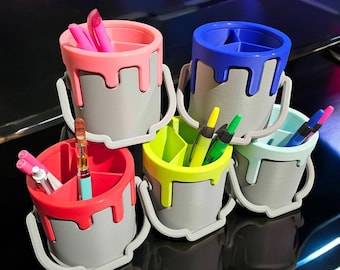 May include: Five paint can shaped desk organizers in different colors, each with a different colored paint drip. The organizers are filled with pens and pencils.