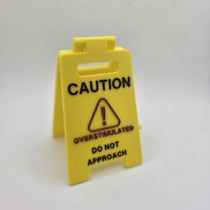 May include: A yellow plastic caution sign with black text that reads "CAUTION" at the top, and "OVERSTIMULATED DO NOT APPROACH" below a red exclamation point in a triangle.