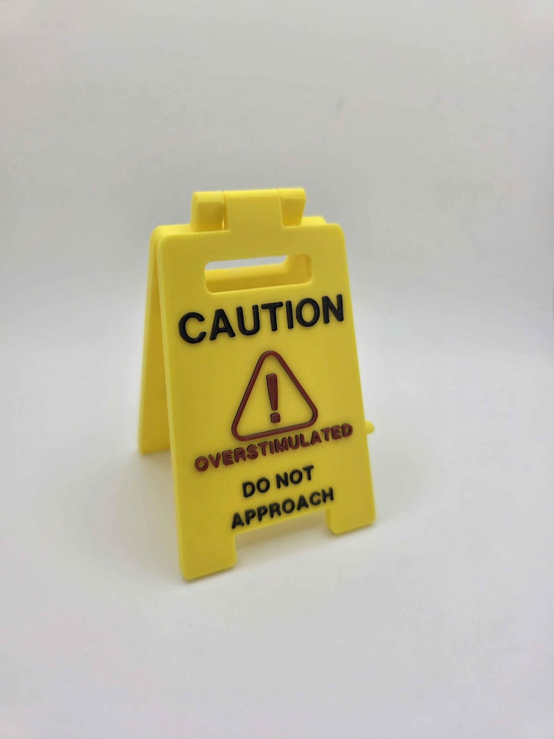 May include: A yellow plastic caution sign with black text that reads "CAUTION" at the top, and "OVERSTIMULATED DO NOT APPROACH" below a red exclamation point in a triangle.